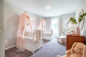 A carpeted nursery with two cradles, a large stuffed animal, and a window overlooking trees.